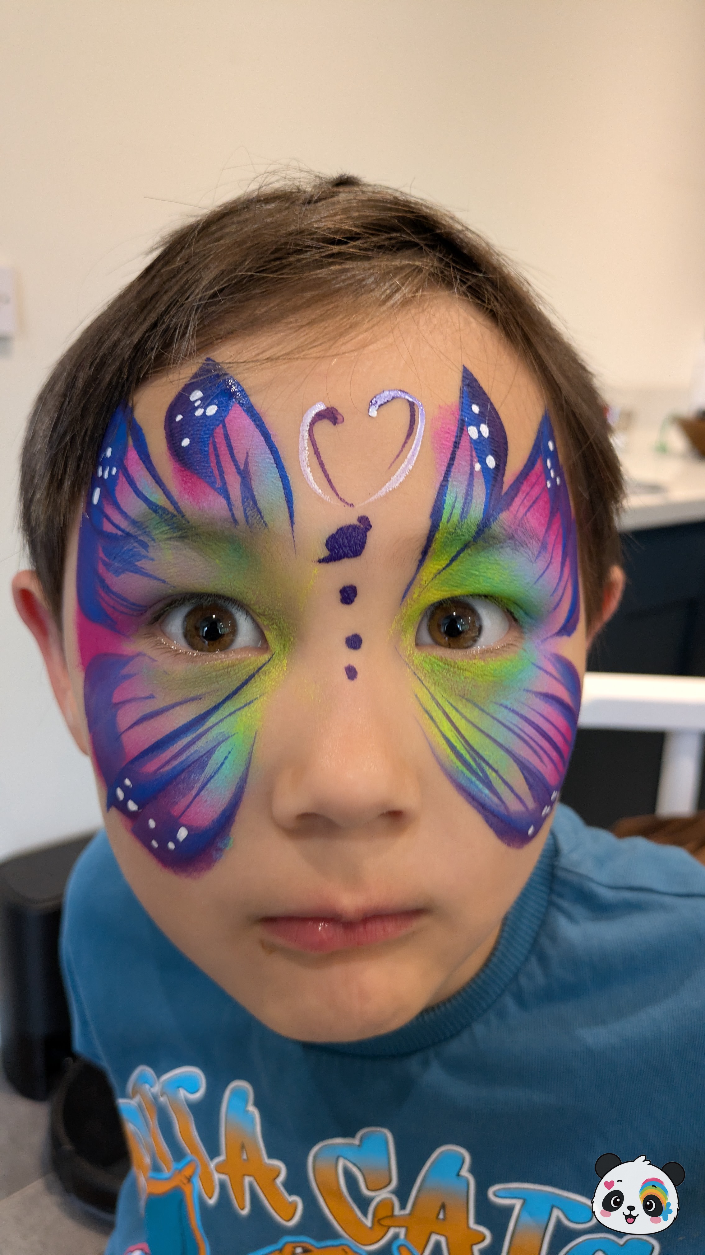 Rainbow butterfly face paint with blue, pink, and yellow wings, a purple heart on the forehead, and white dot accents.