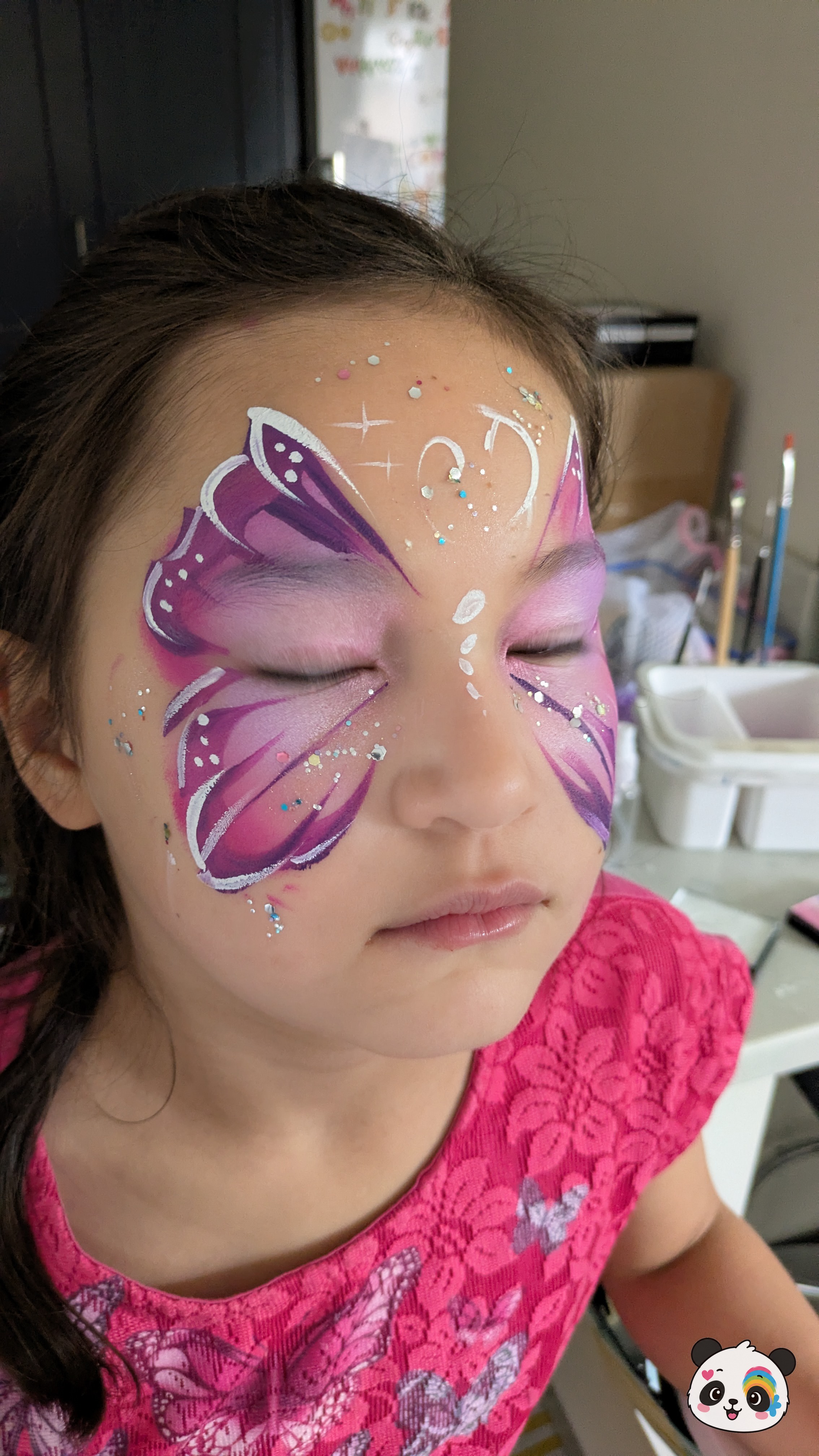 Purple and pink butterfly wing face paint with white highlights and glitter accents across the eyes and forehead.
